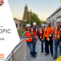 Group of construction workers with orange hi vis vests standing on jobsite with hardhats: Daily Topic Workplace Culture Construction Inclusion Week