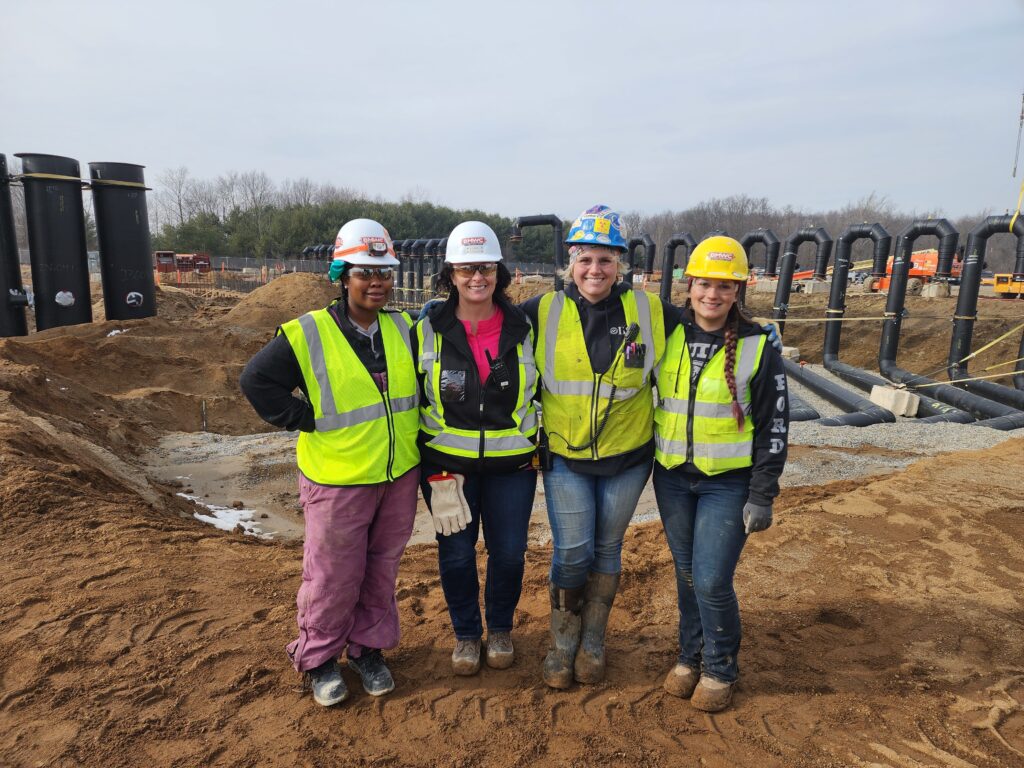 Women celebrating women in construction week - wearing women's construction workwear on job site with candy cane piping in background. 