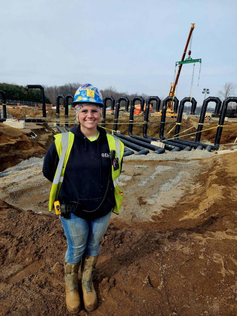 Apprentice and fill-in foreman lauren ozuk wearing womens construction clothes. Crane in background placing candy cane piping system job site. 